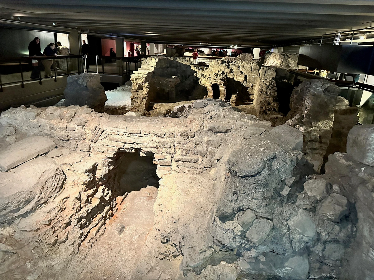 ruins consisting of the bottom sections of stone walls and arches forming passages for (apparently) drainage or an aqueduct. these are enclosed in an underground museum, the walkways and roof of which are visible to the sides.