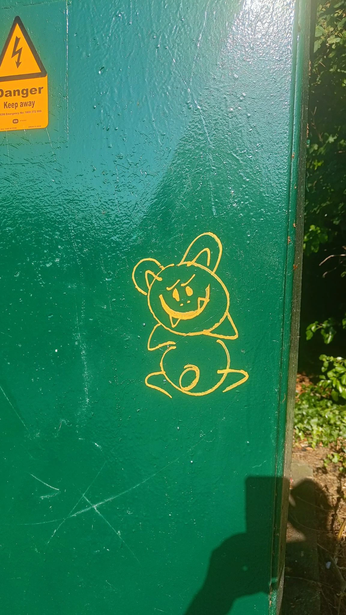 Public electricity box in green with a yellow smiling creature in outline on it