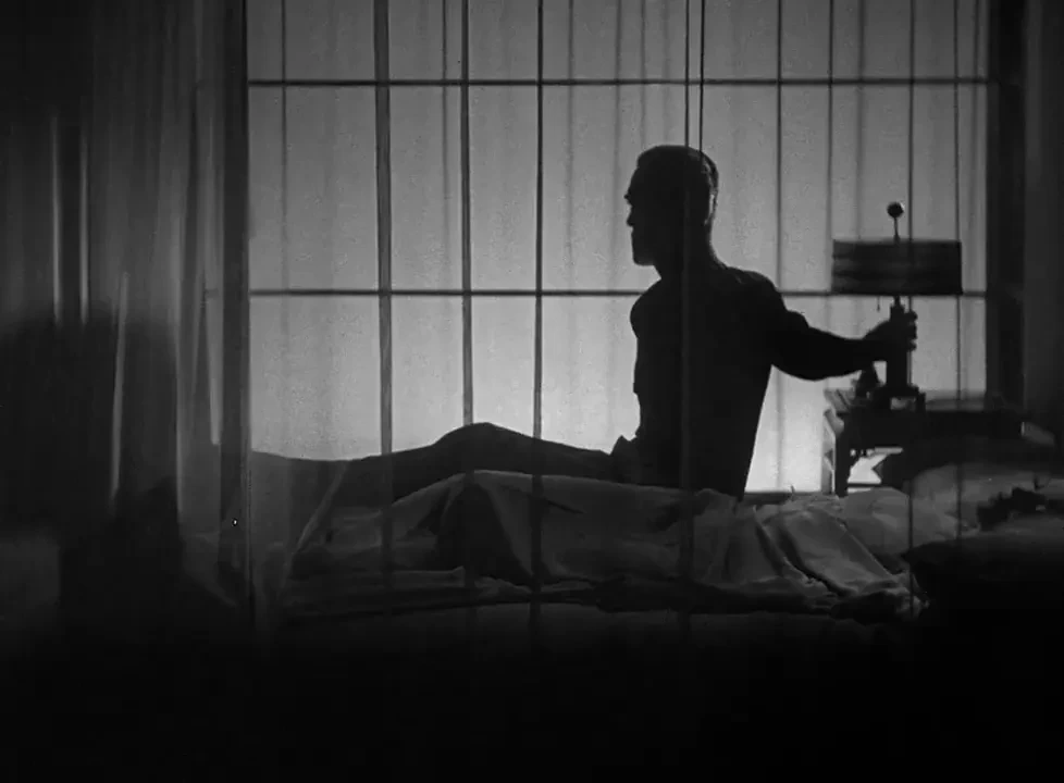 A man sat up in a bed surrounded by thin net curtains is silhouetted by the bedside lamp he just turned on. In the foreground, a young woman with long blonde hair lies on the bed, eyes closed.