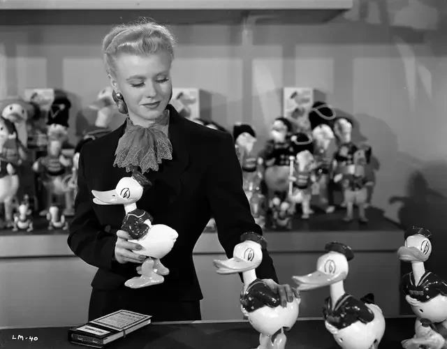 A black & white still image from the 1939 film 'Bachelor Mother'. A young blonde white woman in a dark suit smiles somewhat as she arranges a row of Donald Duck statues on a countertop.