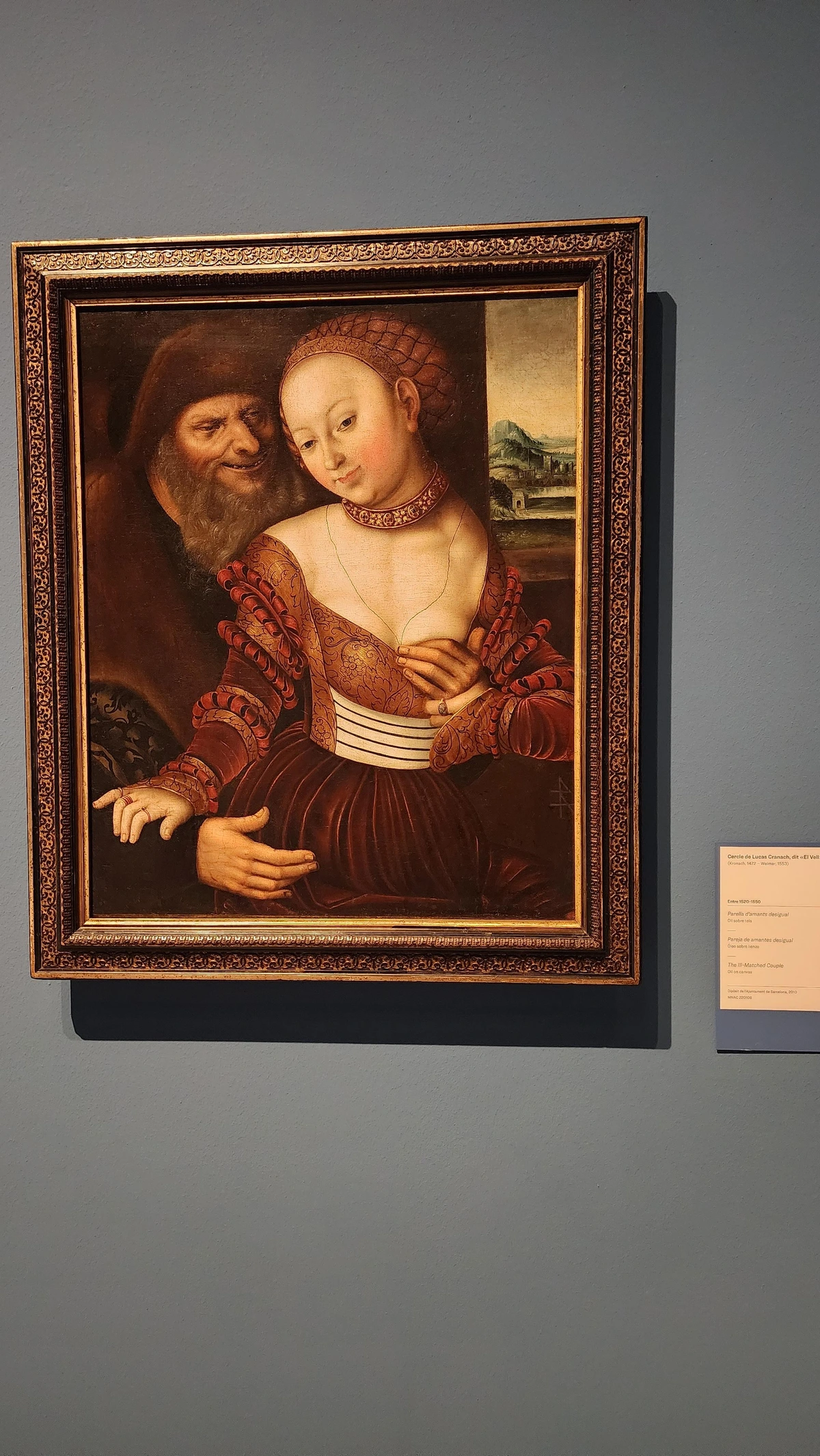A Renaissance oil painting shows two figures in close proximity. A young woman with pale, rosy skin wears an elaborate rust-red brocade dress with puffed sleeves, a netted hair covering, and a jeweled choker. She gazes downward with a faint, ambiguous smile, her hands clasped at her chest. Behind her looms an elderly man with a white beard and dark fur hat, leering toward her with a grasping hand. A landscape with mountains is visible through a window behind them. The contrast between youth and age, beauty and desire, creates an unsettling tension. Attributed to the circle of Lucas Cranach the Elder.