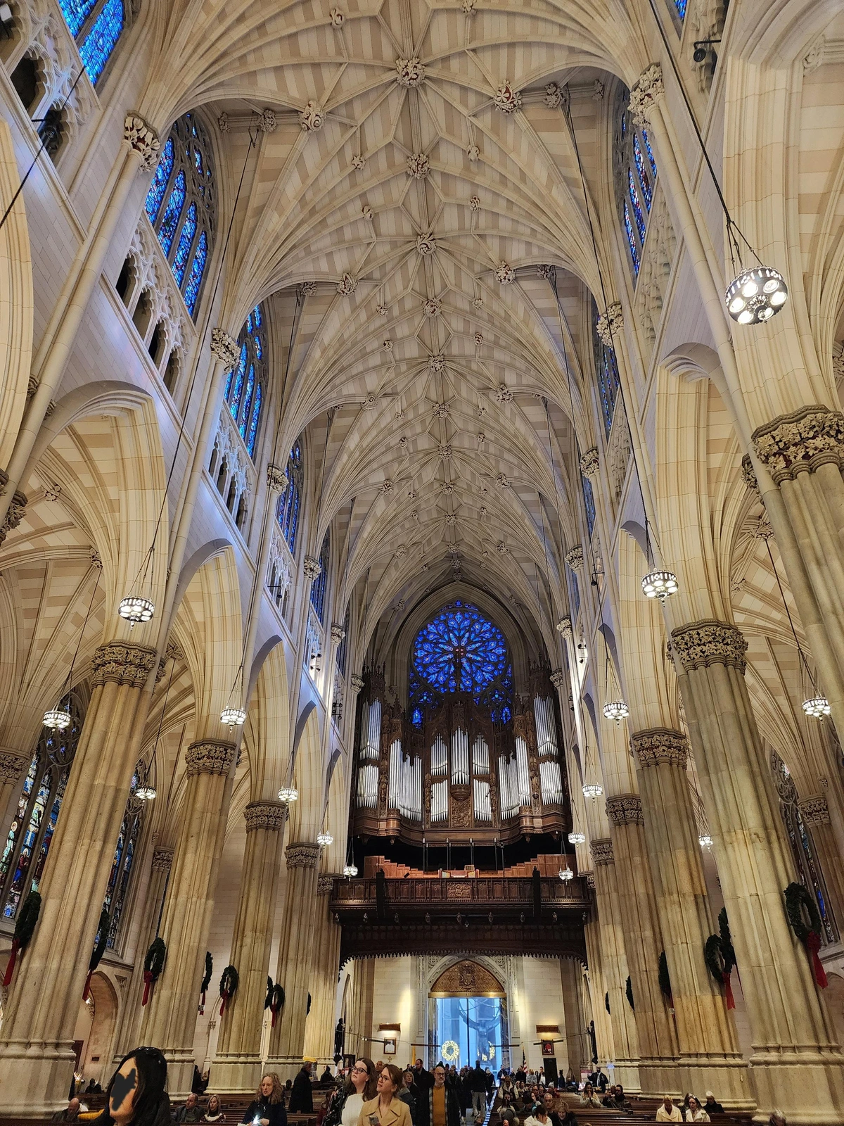 View from the altar to the huge organ above the entrance, showing much of the length of the ribbed vaulted ceiling. above the organ is a  large round blue stained glass window. on either side are columns dividing the main nave from the side naves, and there are people filling the aisle and pews.