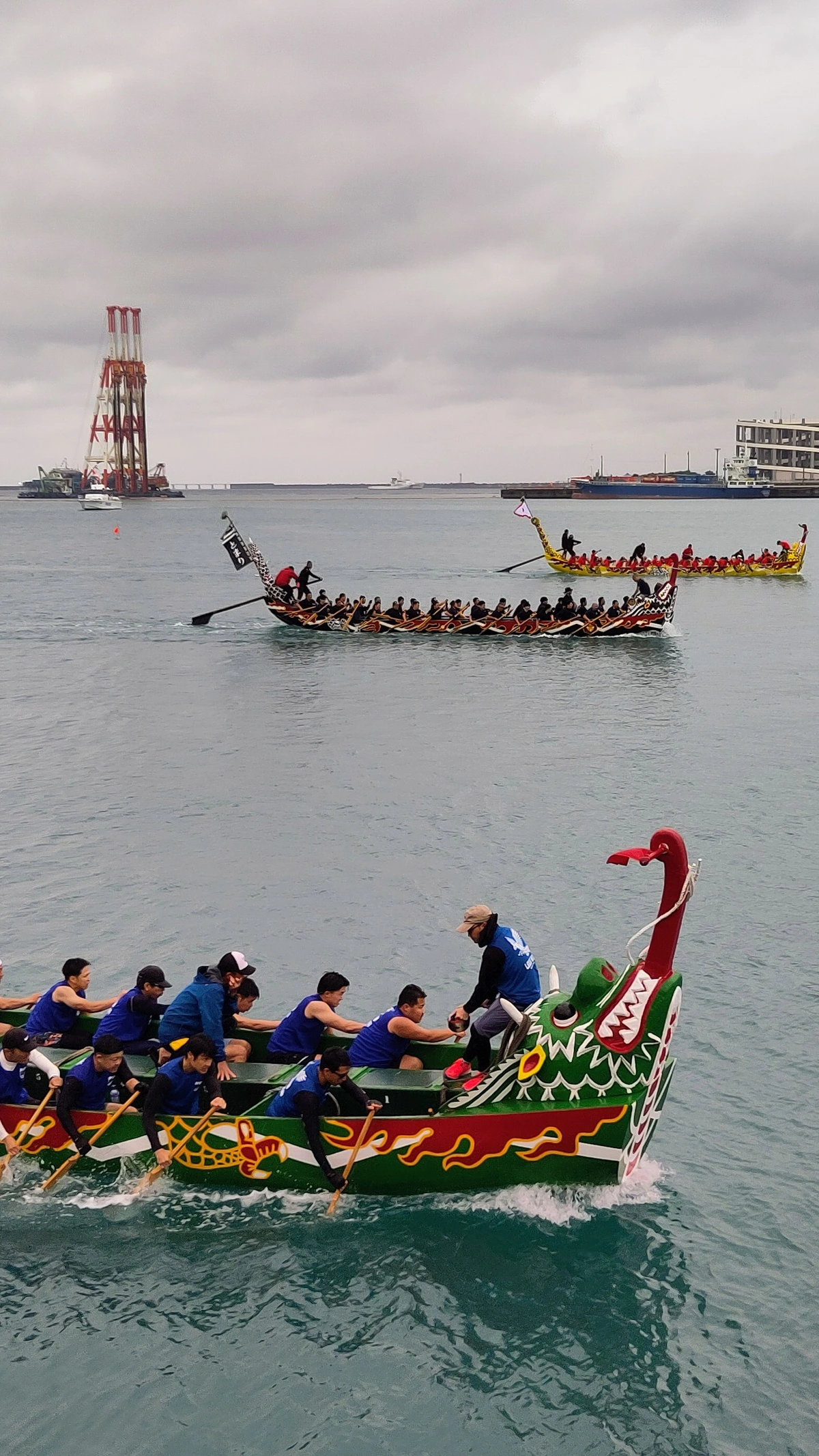 3 Hari boats racing, each with about 20 men paddling in the middle of a port with big ships in the background.