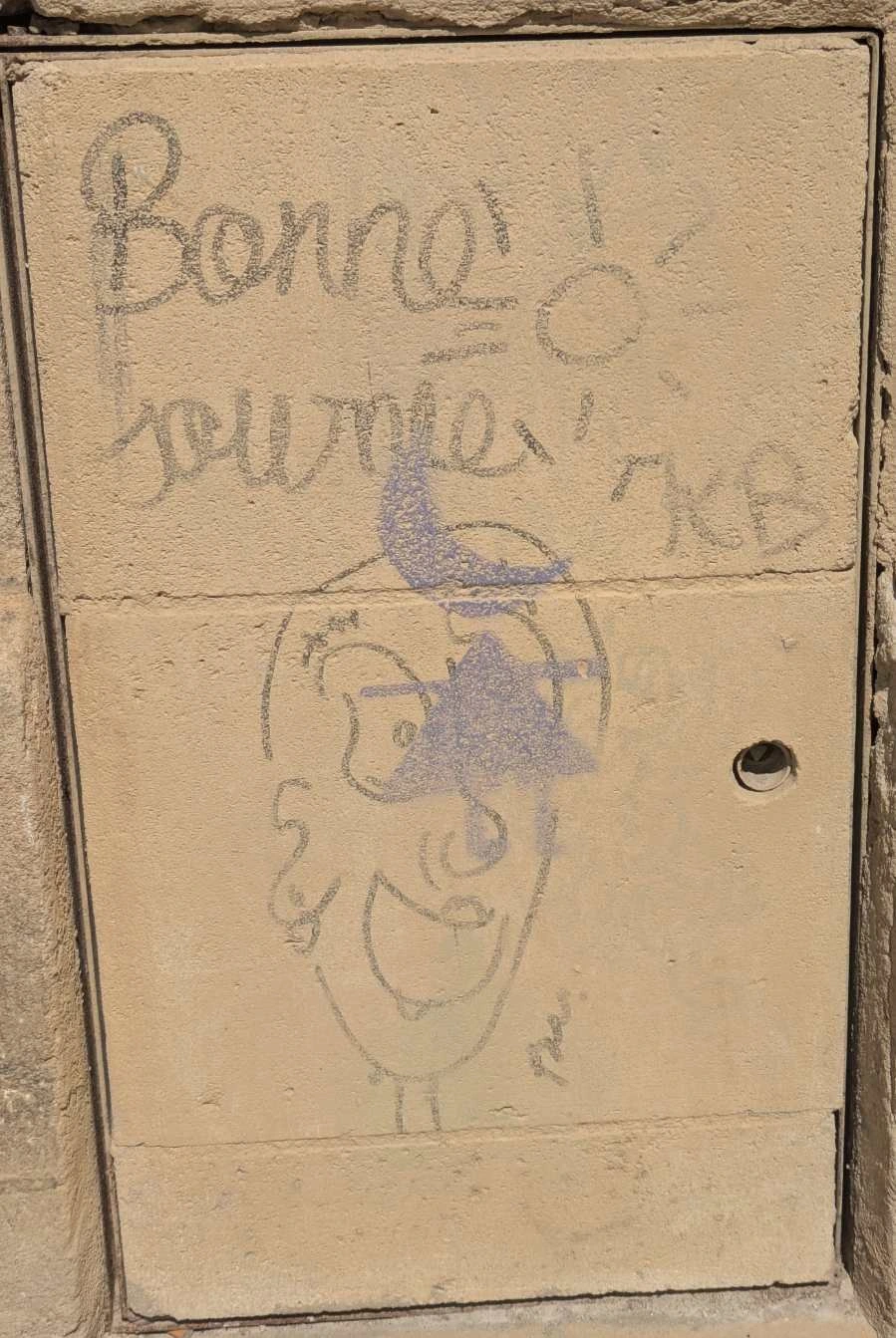 Faded "Bonne Journée" with a happy punk face under it. Written with black pen on light brown stone wall