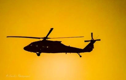 "A Black Hawk helicopter cuts across a sky that looks as if it’s been set on fire by the rising or setting sun. The entire backdrop glows in deep oranges and molten yellows, the kind of light that turns the air itself into a warm haze. Against that brilliance, the helicopter appears as a perfect silhouette—sharp, dark, and unmistakably purposeful.

You can make out the broad main rotor, frozen mid‑spin in the still image, and the smaller tail rotor at the back. The landing gear hangs beneath the fuselage, giving the aircraft a slightly predatory posture, as if it’s ready to drop into action at any moment. The silhouette is crisp enough that every angle and contour reads clearly, even without visible detail. It feels like motion suspended—an instant of power and precision held in the glow of a dramatic sky.

The contrast between the fiery background and the dark aircraft gives the scene a cinematic intensity. It’s a moment that feels both calm and charged, as if the helicopter is moving through the last breath of daylight or the first breath of morning." - Microsoft Copilot