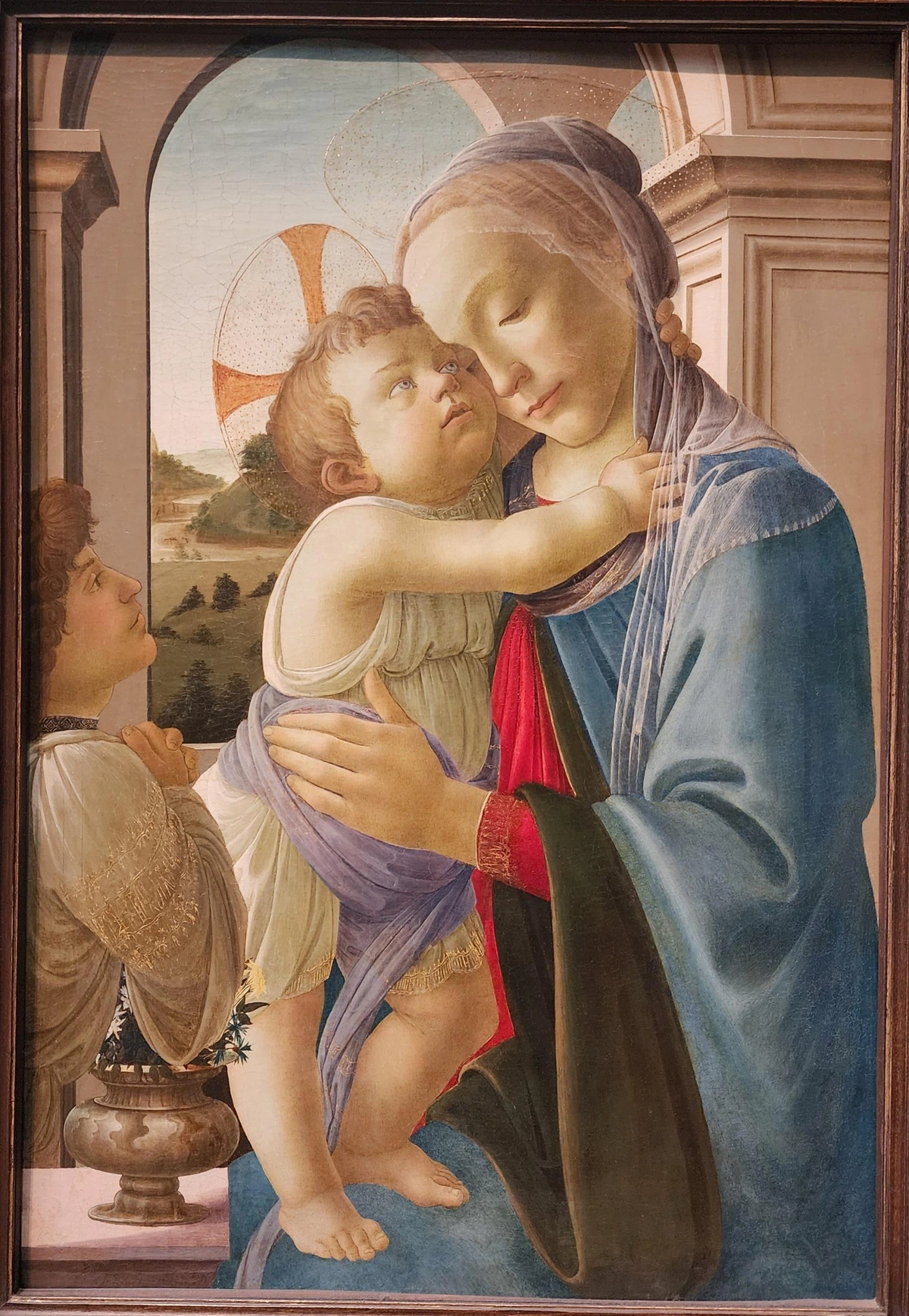 a seated Mary in blue garb and veil holding a baby/toddler Jesus who is standing on her right knee and wrapping his arms around her neck, looking attentively at her. at left is a young man (angel) almost out of frame adoring the child