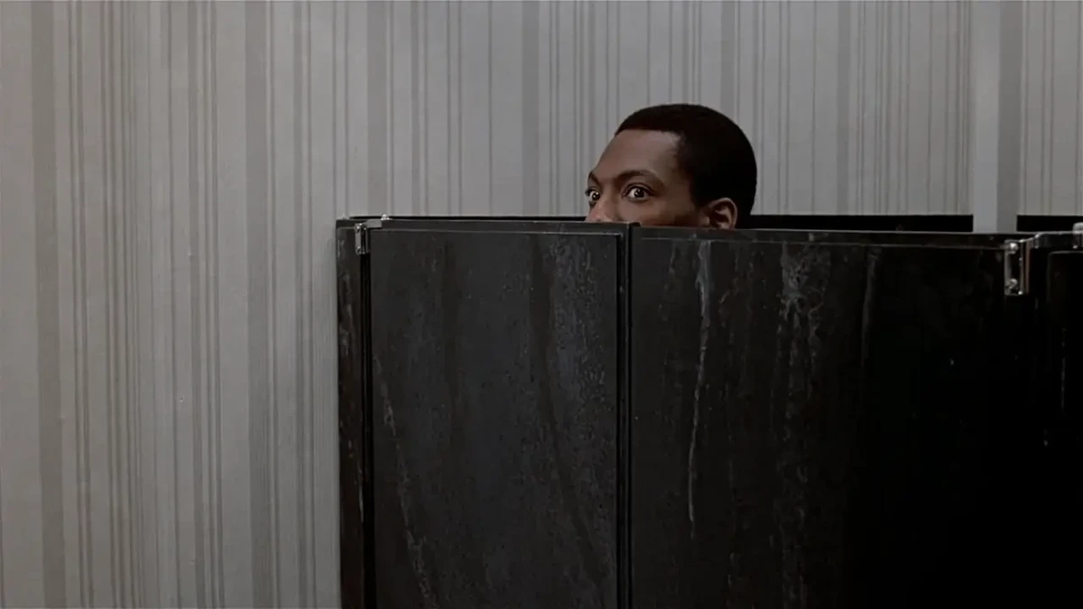 A man, face half-visible, peers out over the marble cubicle door in a high-class toilet
