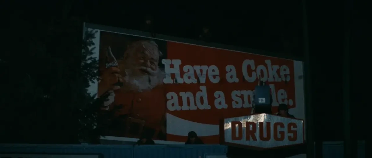 A hoarding with Santa Claus and the slogan "Have a Coke and a smile." Just in front & below, a shop sign says "DRUGS"