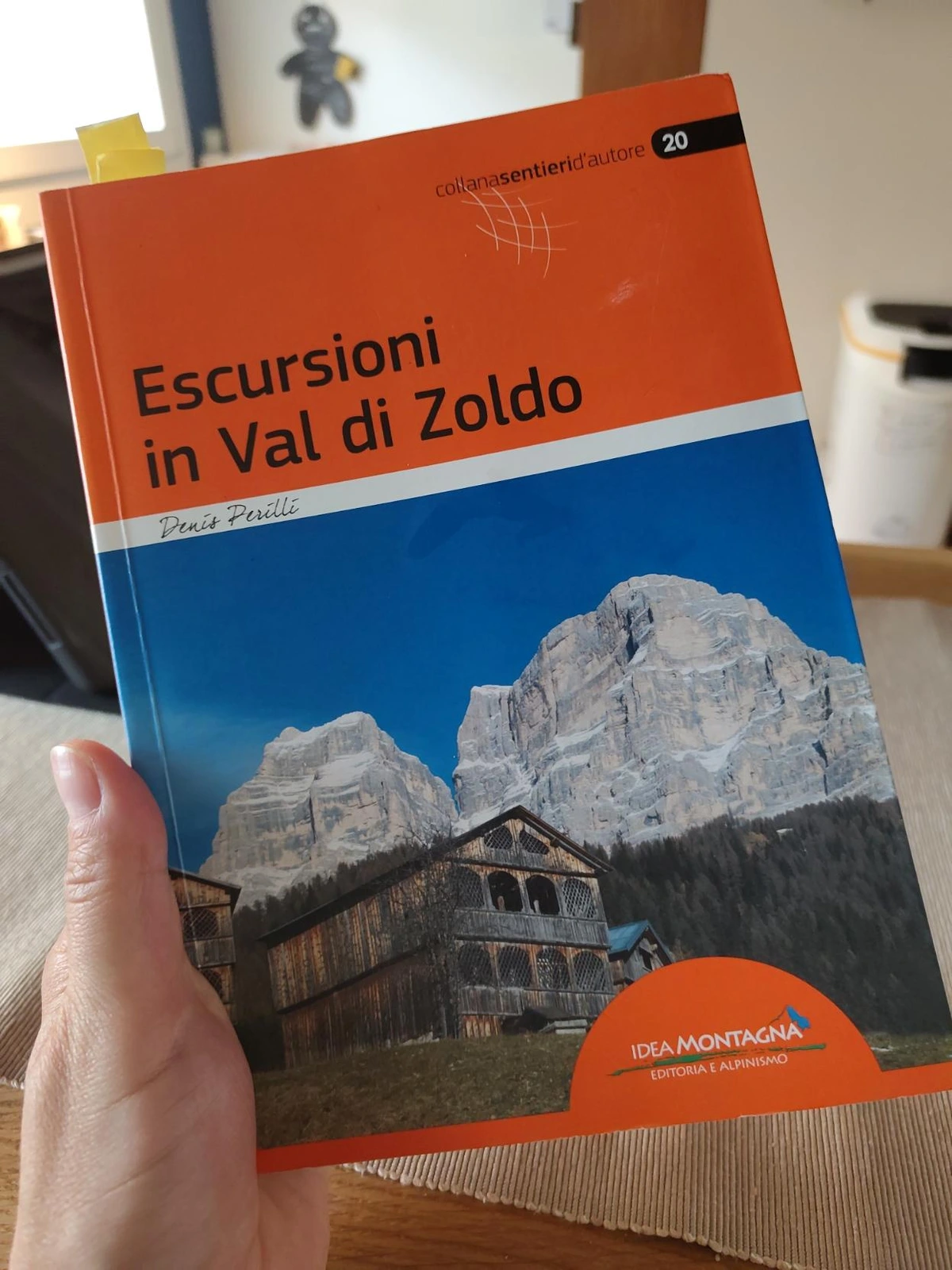 A hand holds an open book titled 'Escursioni in Val di Zoldo' by Denis Perilli, part of the 'collana sentieri d'autore' series. The cover features a striking image of snow-capped mountain peaks (the Pelmo mount) towering over a traditional wooden building with a balcony, set against a backdrop of dense green forests. The book is published by Idea Montagna, as indicated by the logo and text at the bottom right corner, emphasizing its focus on hiking and alpine exploration in the Val di Zoldo region.