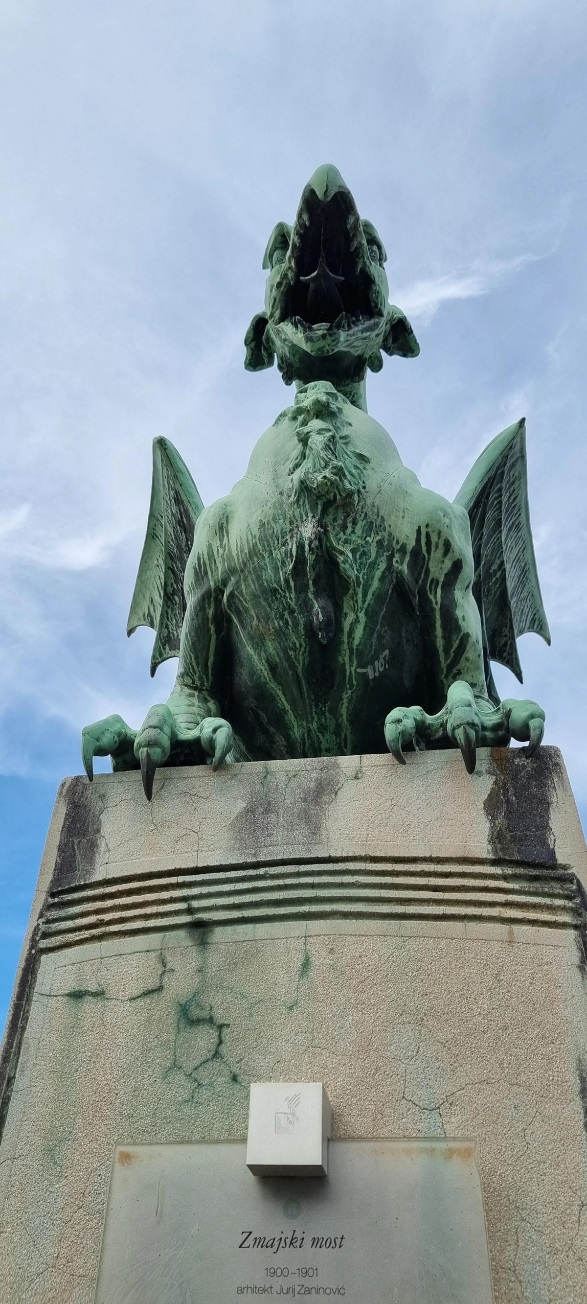 Statue an der Drachenbrücke in Ljubljana: Foto aus der Forschperspektive aufwärts. Auf einem Pfeiler sitzt die Statue eines Drachen, der sein Maul zu einem Schrei öffnet. Durch die Perspektive wirkt der Betrachter dem Drachen unterlegen. Auf dem Pfeiler ist ein Schild angebracht, auf dem steht: "Zmajski most. 1900–1901. Arhitekt Jurj Zaninović"