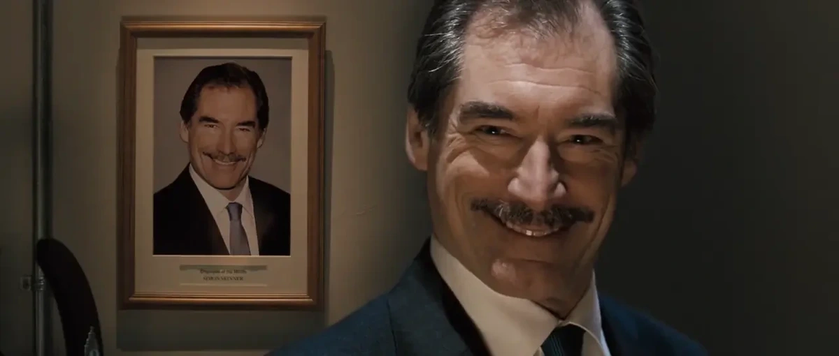 Middle-aged white man with moustache in suit and tie, grinning. On the wall behind him, a photo of him that looks exactly the same.