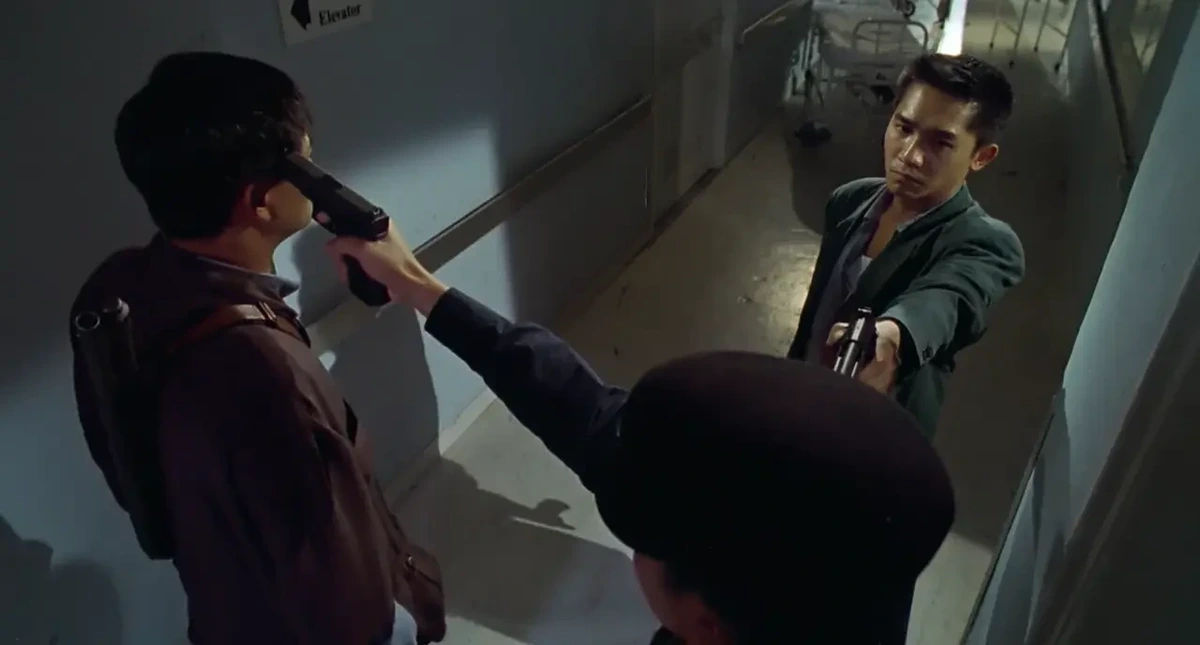 Three men standing in a corridor. Man on right in green jacket is pointing a pistol at the head of man at bottom right in peaked cap, who in turn is pointing a pistol at the head of man on left in brown jacket