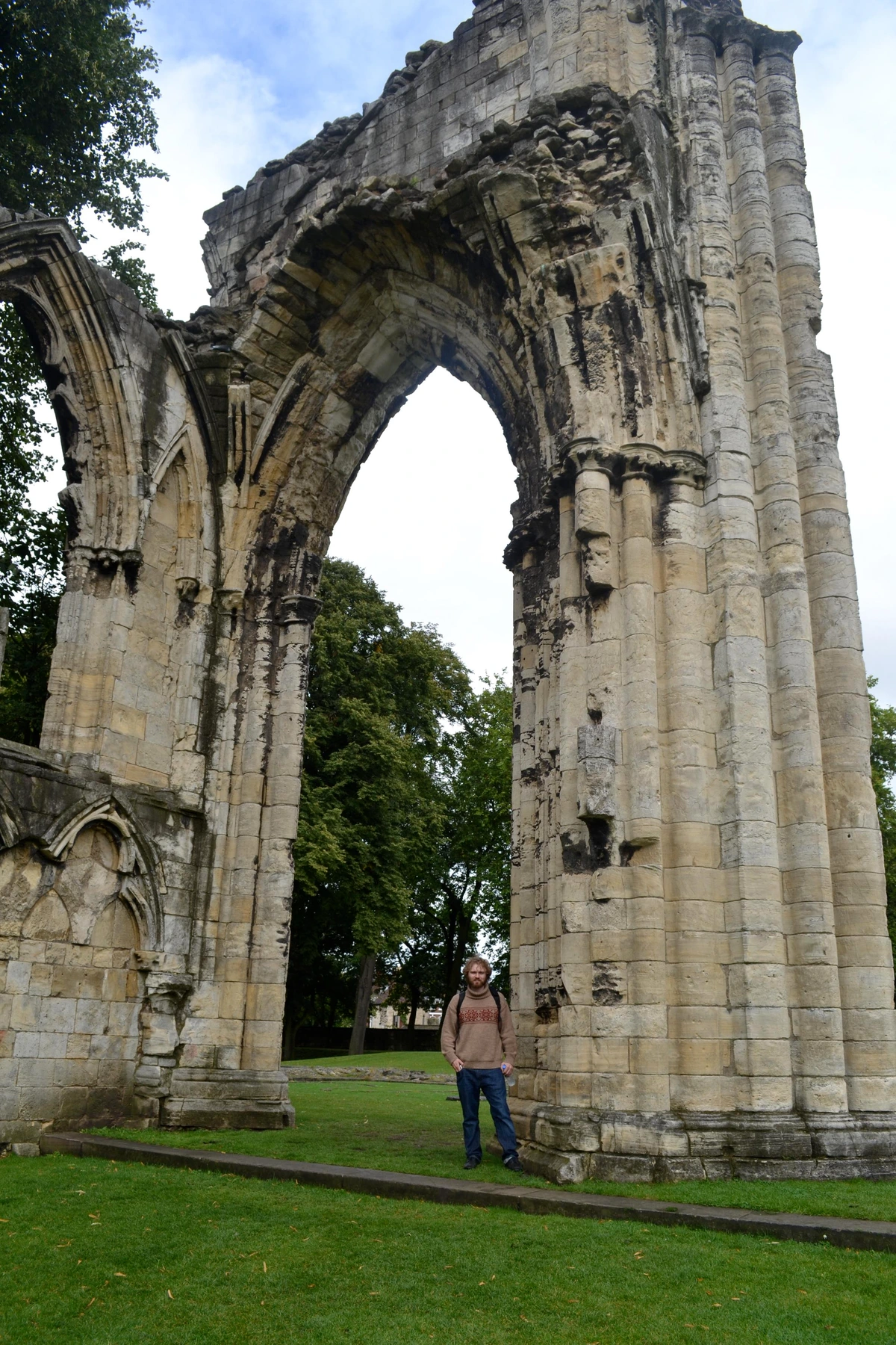 Huge ruined pointed arch of the church entrance in a grassy park. dwarfed at the large base of the arch is a bearded man in a sweater.