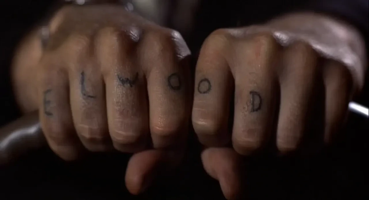 Closeup of two hands gripping the top of a steering wheel. On 6 of the fingers are tattooed one letter per finger: E L W O O D