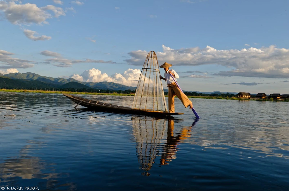 A photograph of one of the fisherman of Inle Lake in Myanmar (aka Burma). The fisherman is in the centre of the frame standing on his small boat. He is holding onto the frame of his conical net in one hand while the other is holding his single oar (his left leg is stretched out along the oar with is foot hooked onto it). He is reflected in the still waters of the lake, as are the clouds in the largely clear blue sky.