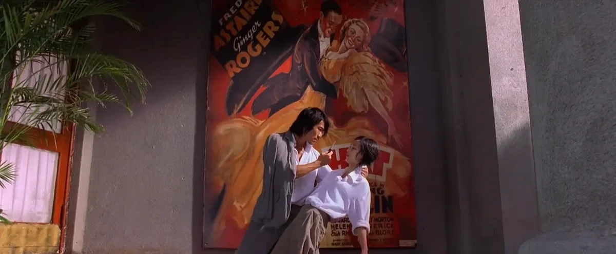 An East Asian man with a light blue jacket draped over his shoulders holds a young east Asian woman in a white blouse leaning away from him. He holds a knife in his other hand close to her face. Behind them is a large painted film poster featuring Fred Astaire & Ginger Rogers dancing. The poses of the people and the movie stars parallel each other.