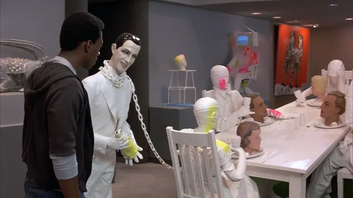 A man at left looks at an art piece consisting of a mannequin in white and a dinner table with mannequins seated at it, with mannequin heads on the plates.