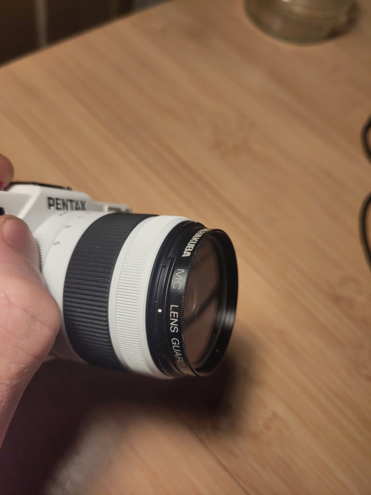 Closeup on a camera lens, attached to the camera. The lens has a lens guard on it. The guard is branded Hakuba, and is labelled as an MC Lens Guard. 

The camera is a blue and white Pentax Q10. The background is a wooden table. 