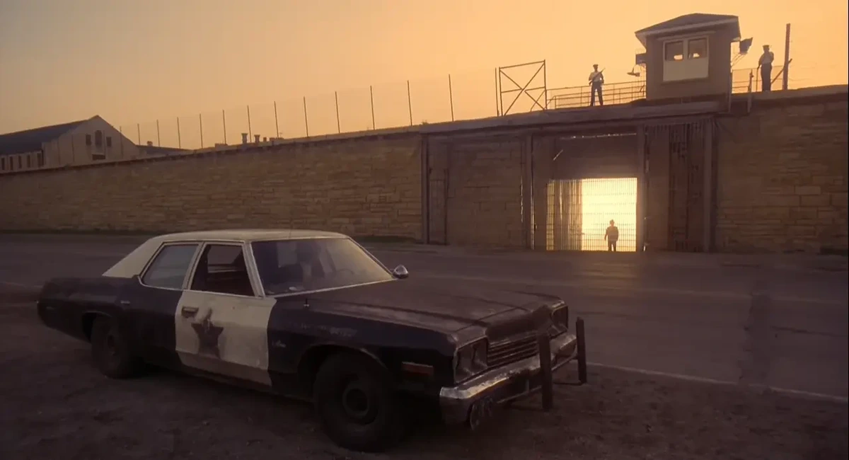 A dirty former police car is parked on the near side of a road. On the other side is a long wall with wire & guards on top. A gate is open and early morning sunlight shines through, silhouetting a lone figure.