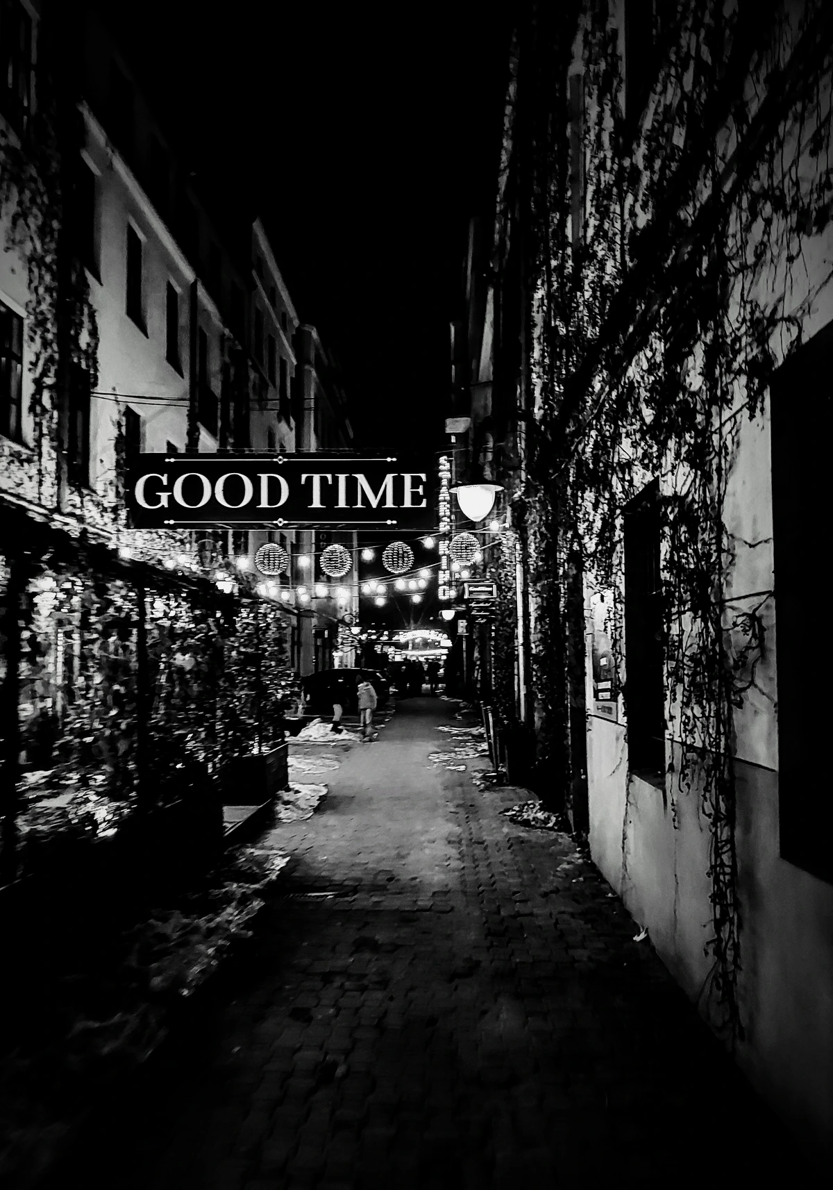 backyard of townhouses, in the dark, snow. obvious repurposing happening as restaurant can be seen to left, hanging lights ahead, ivy-covered wall to right. a huge sign saying "good time" is hanging over the yard