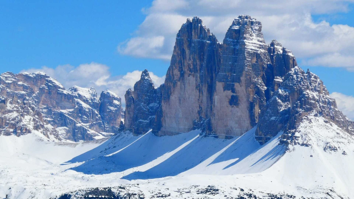 Una spettacolare vista delle Tre Cime di Lavaredo, con le loro imponenti guglie rocciose che si ergono verso il cielo azzurro e limpido. Le cime sono parzialmente coperte di neve, e la luce del sole accentua le sfumature delle rocce, creando un contrasto con il bianco della neve sottostante. Il paesaggio trasmette un senso di maestosità e bellezza naturale, con le montagne che dominano l'orizzonte
