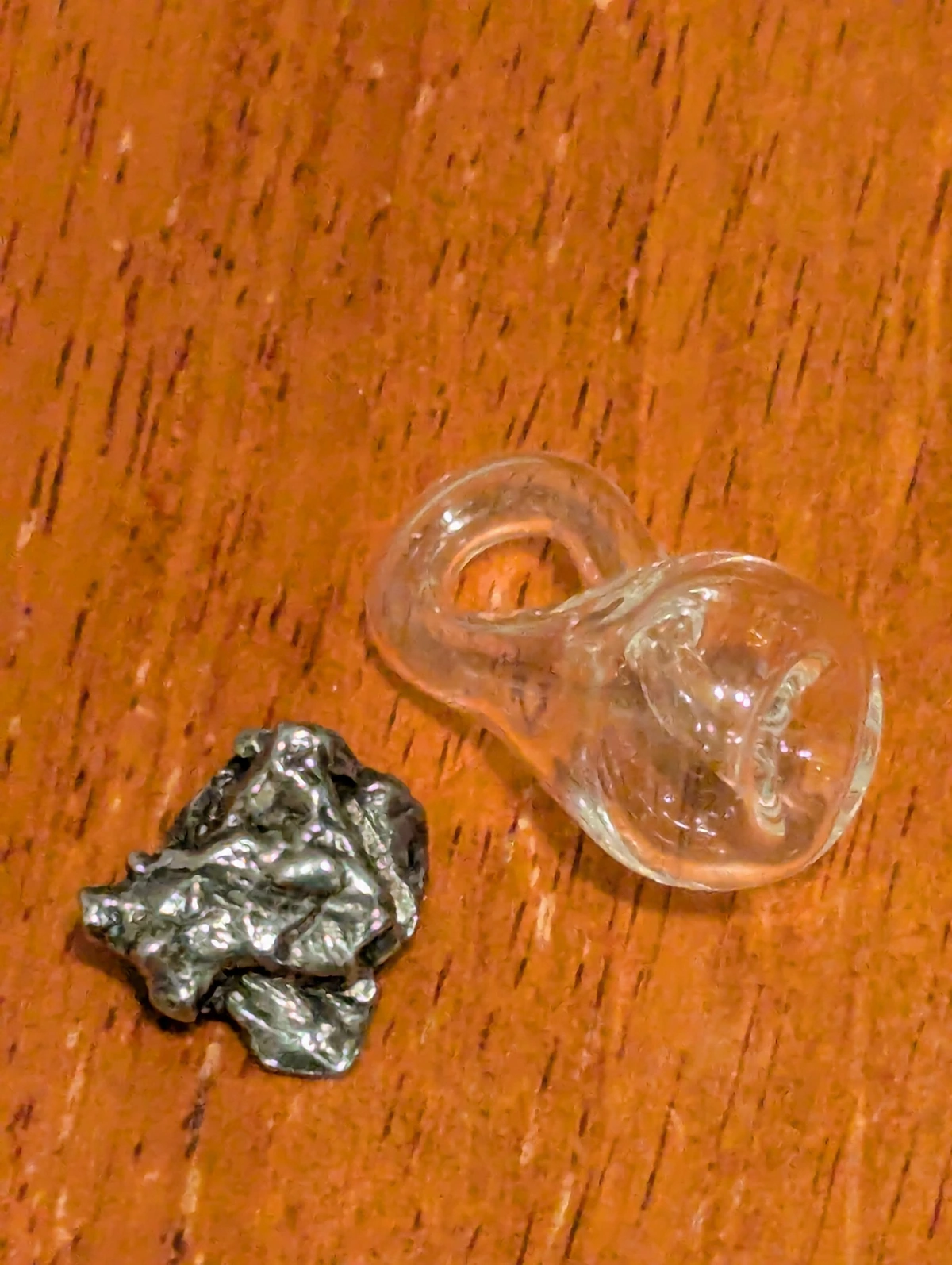 A small irregular shiny metal pebble next to a glass Klein bottle on a wooden surface