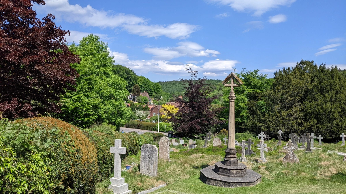A churchyard in a rural English village on a sunny day. A cross monument is in the foreground with many gravestones surrounded by thick bushes and trees. Part of a road and some houses are visible beyond the churchyard. In the distance is a hill covered in forest.