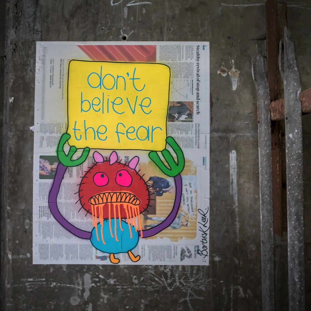 A scaremongering newspaper article is attached to a wall with a brightly coloured child-like drawing of a drooling creature holding a sign saying "don't believe the fear" painted over it