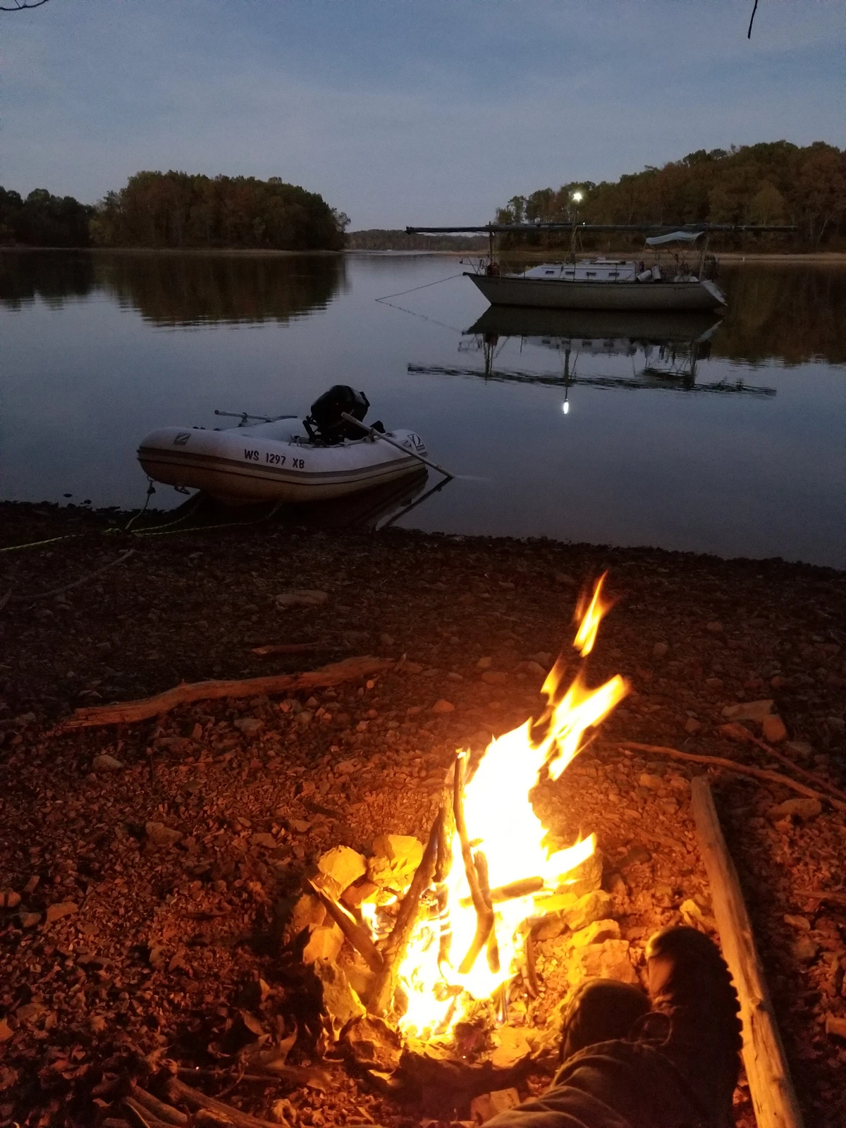 Feet and a campfire in the foreground, with a dinghy pulled up on shore and a sailboat with the mast down sits at anchor.
